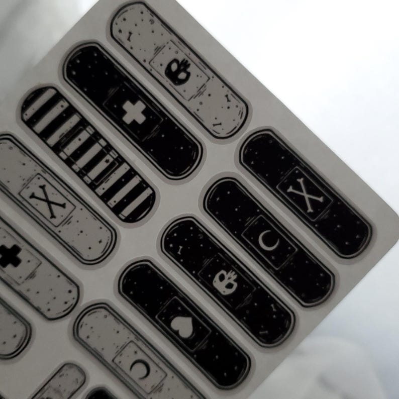 May include: A sheet of black and white adhesive bandages with various designs, including a cross, a crescent moon, a heart, a crown, and a skull and crossbones.