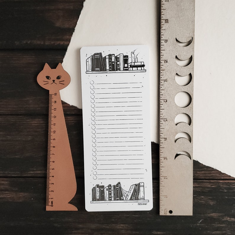 K&ouml;nnte beinhalten: Eine wei&szlig;e Checkliste mit einer schwarz-wei&szlig;en Illustration eines B&uuml;cherregals, das oben und unten mit B&uuml;chern gef&uuml;llt ist. Die Checkliste hat 30 leere Zeilen und 30 Kreise zum Abhaken. Der Text "PAPER & MINT" befindet sich am unteren Rand der Checkliste.