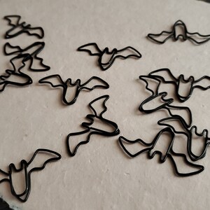 May include: A collection of black metal bat-shaped paper clips. The clips are arranged in a random pattern on a light beige surface.