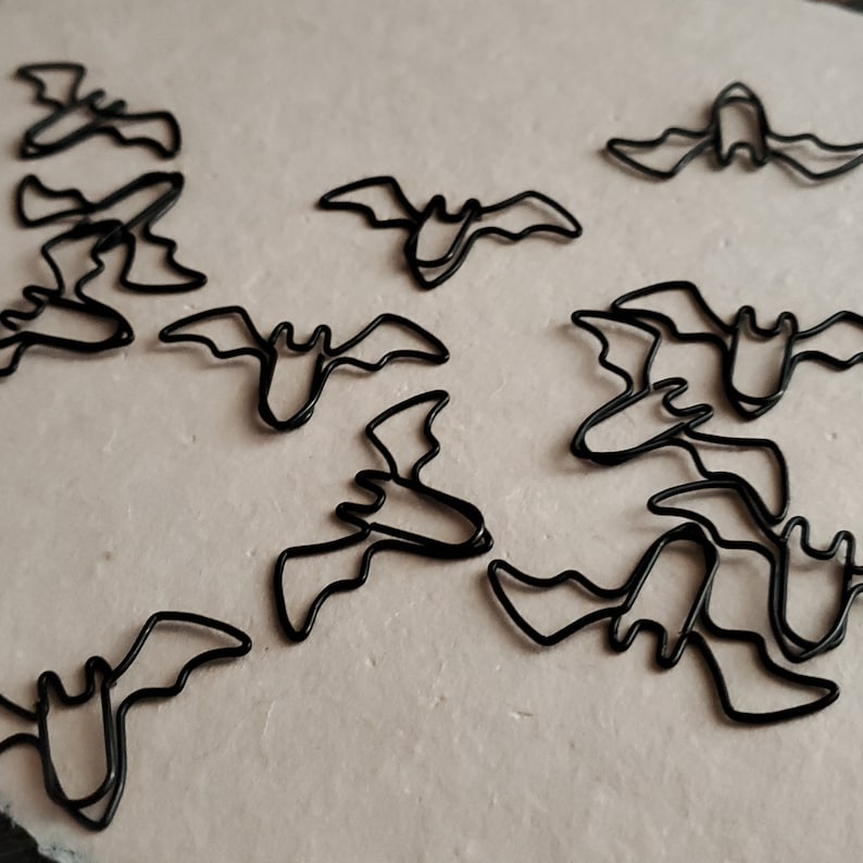 May include: A collection of black metal bat-shaped paper clips. The clips are arranged in a random pattern on a light beige surface.