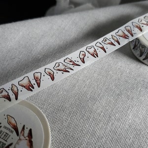 May include: White washi tape with a repeating pattern of brown teeth. The tape is rolled up and the label reads "Bloody Teeth Washi"