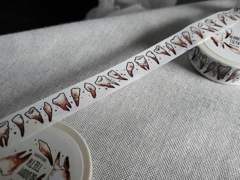 May include: White washi tape with a repeating pattern of brown teeth. The tape is rolled up and the label reads "Bloody Teeth Washi"