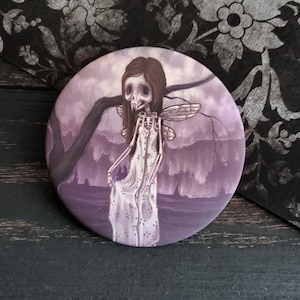 May include: A round mirror with a purple and white image of a skeletal fairy with wings. The fairy is standing in a purple forest with a large tree branch behind her.