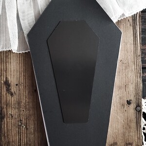 May include: A black coffin-shaped magnet with a rectangular inset, set against a dark background. The magnet is made of a matte material. The background is a mix of wood and white lace.