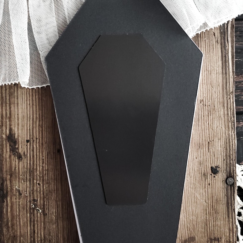 May include: A black coffin-shaped magnet with a rectangular inset, set against a dark background. The magnet is made of a matte material. The background is a mix of wood and white lace.