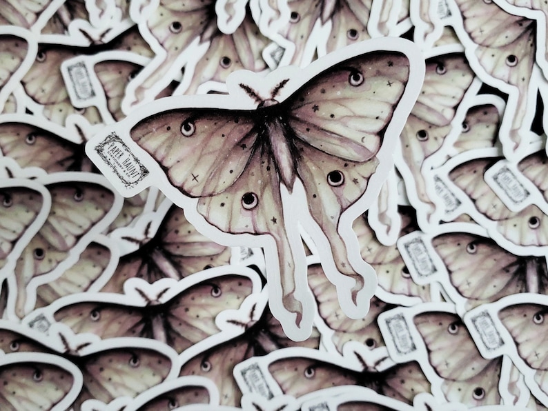 May include: A close-up of a pile of stickers featuring a detailed illustration of a moth with large, iridescent wings and a celestial design. The moth has a delicate, almost translucent appearance, with a subtle pink and beige color scheme. The stickers are printed with the text "Paper Dainty" in a vintage font.
