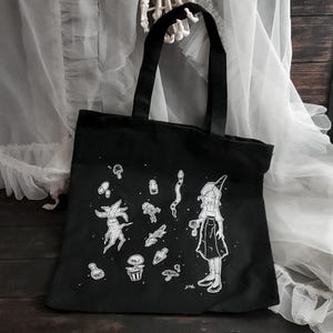K&ouml;nnte beinhalten: Schwarze Canvas-Tragetasche mit einer wei&szlig;en Illustration einer Hexe mit einem spitzen Hut, die eine Blume h&auml;lt, umgeben von verschiedenen magischen Gegenst&auml;nden wie Pilzen, Pflanzen und Tr&auml;nken. Der Text "STK." befindet sich am unteren Rand des Designs.
