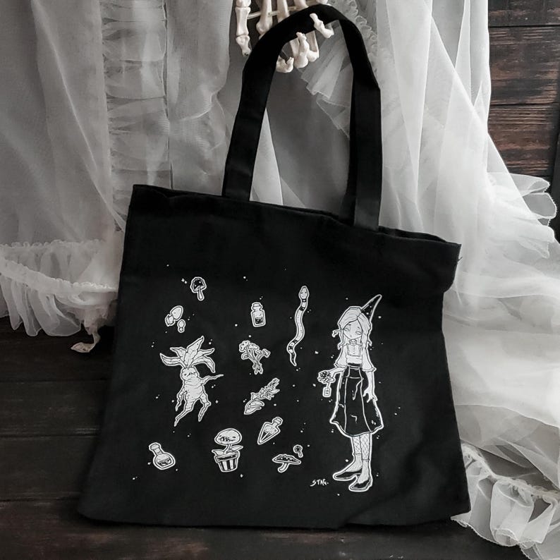 K&ouml;nnte beinhalten: Schwarze Canvas-Tragetasche mit einer wei&szlig;en Illustration einer Hexe mit einem spitzen Hut, die eine Blume h&auml;lt, umgeben von verschiedenen magischen Gegenst&auml;nden wie Pilzen, Pflanzen und Tr&auml;nken. Der Text "STK." befindet sich am unteren Rand des Designs.