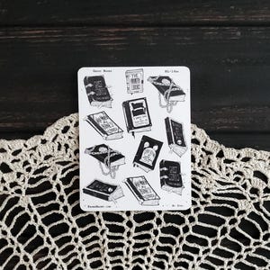 May include: A sheet of black and white stickers featuring illustrations of spooky books with titles like "Ghost Stories", "The Haunted Library", and "Within My Bones". The stickers are on a white background and have a vintage aesthetic.