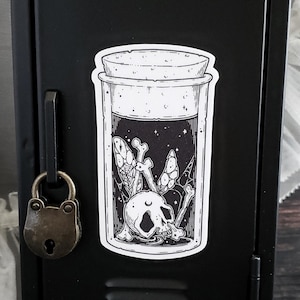 May include: A black and white sticker featuring a jar with a cork lid. Inside the jar is a skull, bones, and a starry night design. The sticker is affixed to a black metal surface, next to a small padlock.