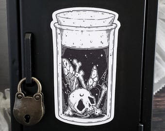 Peut inclure: Un autocollant noir et blanc représentant un bocal avec un couvercle en liège. À l'intérieur du bocal se trouvent un crâne, des os et un motif de nuit étoilée. L'autocollant est sur une surface métallique noire, à côté d'un petit cadenas.