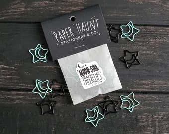 May include: A set of black and light blue star-shaped paper clips with a crescent moon design. The paper clips are packaged in a white paper bag with the text "Paper Haunt Stationery & Co." and "Moon-Star Paperclips" printed on it.