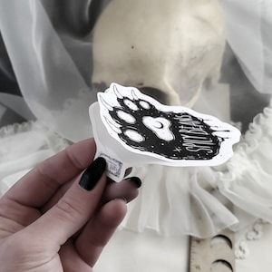 Puede incluir: Una pegatina en blanco y negro con una huella de pata estilizada y el texto "Spicy Beans". La pegatina se sostiene en una mano con esmalte de u&ntilde;as negro. El fondo incluye una calavera y tela blanca.
