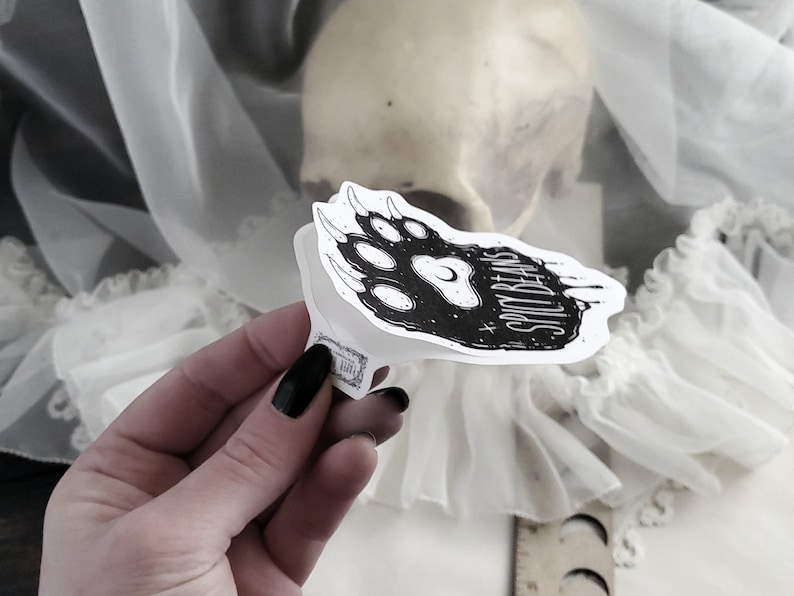 Puede incluir: Una pegatina en blanco y negro con una huella de pata estilizada y el texto "Spicy Beans". La pegatina se sostiene en una mano con esmalte de u&ntilde;as negro. El fondo incluye una calavera y tela blanca.
