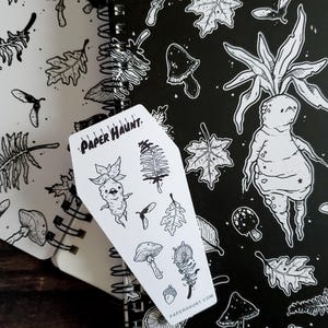 May include: A white sticker sheet with black line drawings of a mushroom, a fern, a leaf, a creature with a mushroom head, and the text "PAPER HAUNT". The sticker sheet is on top of a black and white notebook with a repeating pattern of mushrooms, leaves, and a creature with a mushroom head.