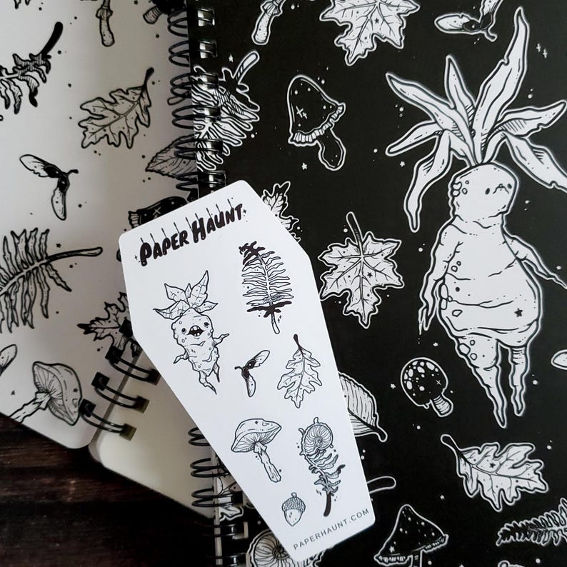 May include: A white sticker sheet with black line drawings of a mushroom, a fern, a leaf, a creature with a mushroom head, and the text "PAPER HAUNT". The sticker sheet is on top of a black and white notebook with a repeating pattern of mushrooms, leaves, and a creature with a mushroom head.