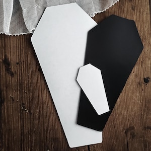 May include: Three coffin-shaped paper cutouts in black and white, arranged on a wooden surface. The largest is white, the next is black, and a smaller white one is in front. A lace fabric is in the background.