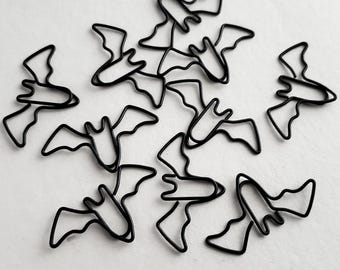 Könnte beinhalten: Eine Ansammlung von zehn schwarzen Büroklammern aus Metall, die jeweils wie eine stilisierte Fledermaus mit ausgebreiteten Flügeln geformt sind. Die Büroklammern sind verstreut angeordnet und zeigen die dünne Drahtkonstruktion sowie die markante Fledermaussilhouette.