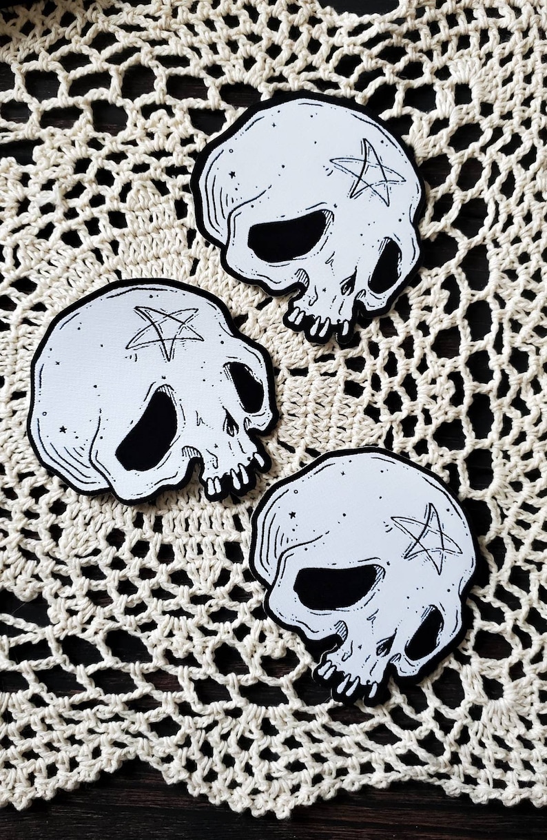 Op de afbeelding: Drie witte schedelvormige patches met zwarte contouren en een pentagram-ontwerp op elk. De patches bevinden zich op een wit gehaakt doily.