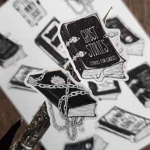 May include: Two black and white illustrated stickers of books. One book is titled "Ghost Stories" with the subtitle "Stories for Ghosts". The other book is chained shut and has the word "Haunted" on the cover.