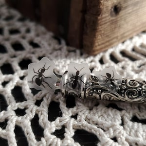 May include: Three clear plastic stickers with black ant silhouettes. The stickers are on a silver, ornate, metal surface.