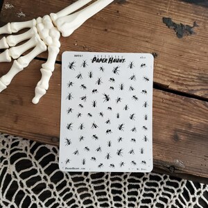 May include: A sheet of black ant stickers with the text "ANTS! PAPER HAUNT. A120" printed on it. The sheet is on a wooden surface.