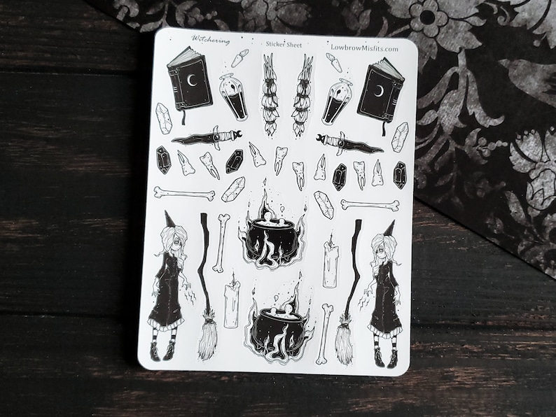 May include: A black and white sticker sheet with a witching theme. The sheet includes a witch, a cauldron, a book, a dagger, bones, crystals, candles, and a broom. The text "Witching" and "Sticker Sheet" are at the top of the sheet. The text "LowbrowMisfits.com" is at the top right of the sheet.
