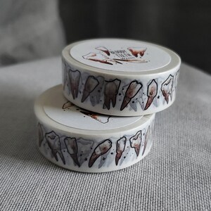May include: Two rolls of washi tape with a white background and a pattern of brown teeth. The text "Bloody Teeth" is printed on the top roll.