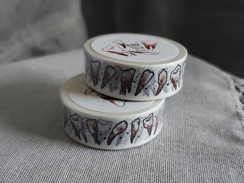 May include: Two rolls of washi tape with a white background and a pattern of brown teeth. The text "Bloody Teeth" is printed on the top roll.
