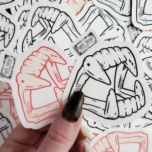May include: A set of stickers featuring a black and white illustration of a set of fangs. The stickers are on a white background and have a red outline. The stickers are from the company "Paper Haunt".