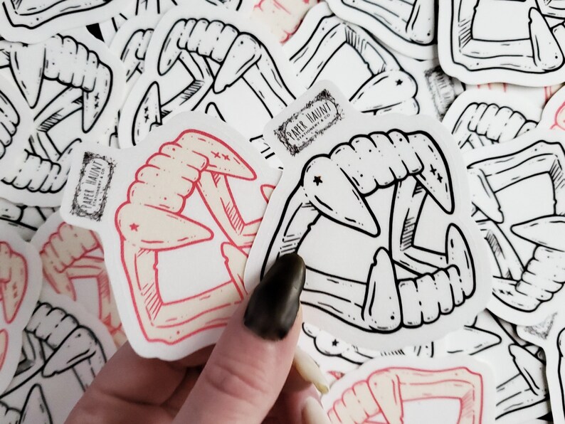May include: A set of stickers featuring a black and white illustration of a set of fangs. The stickers are on a white background and have a red outline. The stickers are from the company "Paper Haunt".