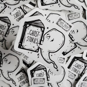 May include: A black and white sticker featuring a ghost holding a book with the text "Ghost Stories" on the cover. The sticker also includes the text "Paper Haunt" and stars.