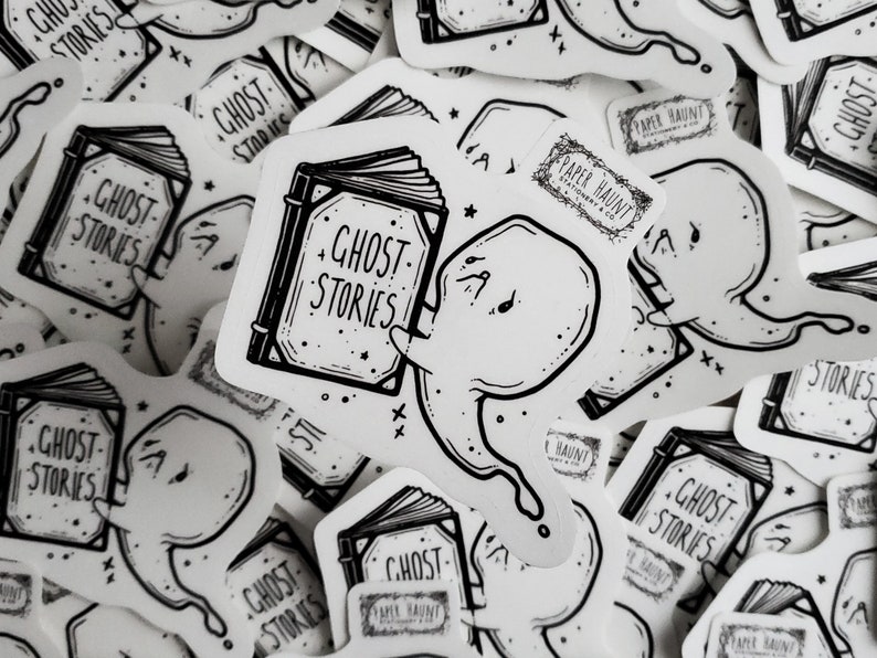 May include: A black and white sticker featuring a ghost holding a book with the text "Ghost Stories" on the cover. The sticker also includes the text "Paper Haunt" and stars.