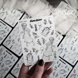 May include: A sheet of black and white stickers featuring a spooky garden theme. The stickers include a girl in a long dress, a tombstone, flowers, and ghosts. The sheet is labeled "Paper Haunt" and "Haunted Garden".