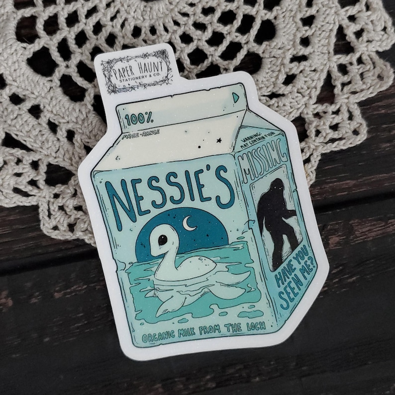 K&ouml;nnte beinhalten: Eine Cartoon-Illustration eines Nessie-&auml;hnlichen Wesens, das in einem blauen See schwimmt. Die Illustration befindet sich auf einem wei&szlig;en Karton mit dem Text "Nessie's Organic Milk from the Loch" und einem Warnhinweis, dass die Milch "Figmente" enthalten kann.