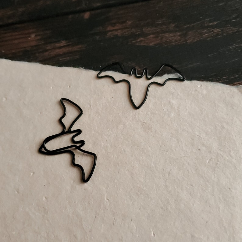 May include: Two black metal bat-shaped paper clips. The clips are on a white surface.
