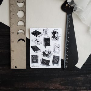 May include: A sheet of black and white stickers featuring illustrations of books with titles like "The Unwritten Library" and "100 Rules for Knowing the Dead".