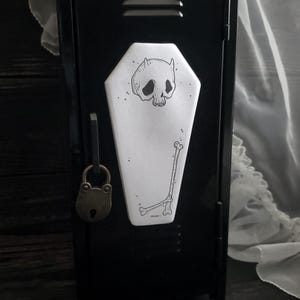 May include: A black metal locker with a coffin-shaped sticker featuring a skull with horns and a bone illustration. The locker has a brass padlock and a latch. The sticker is white with black ink.