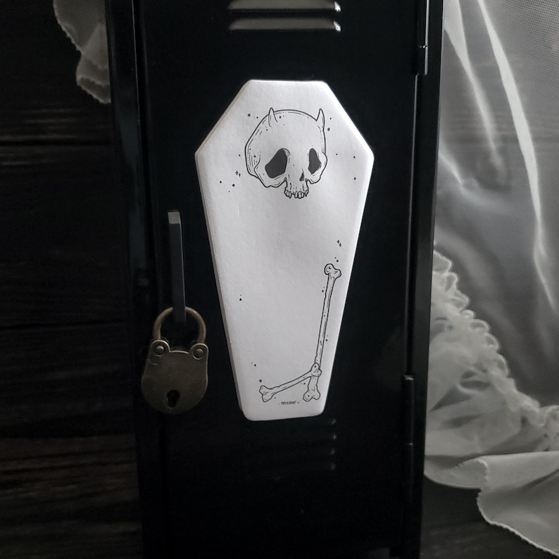 May include: A black metal locker with a coffin-shaped sticker featuring a skull with horns and a bone illustration. The locker has a brass padlock and a latch. The sticker is white with black ink.