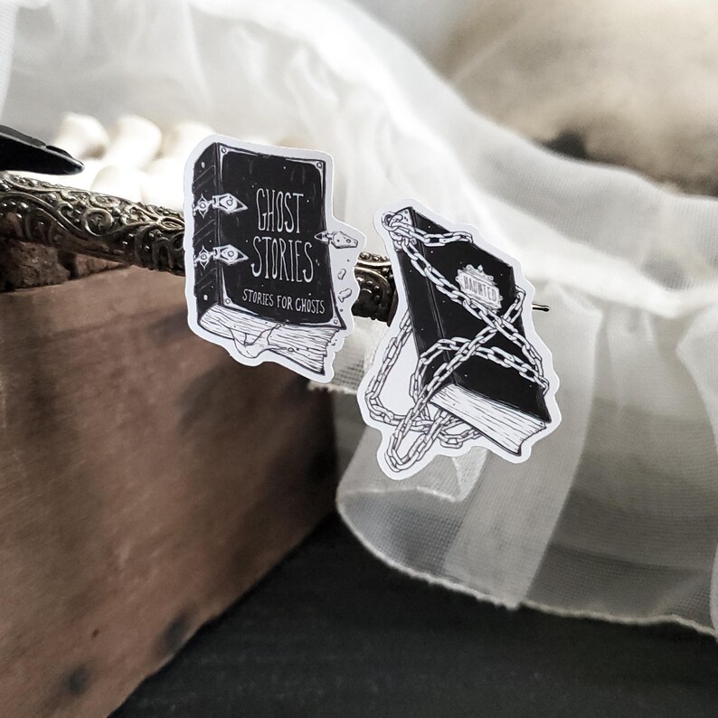May include: Two black and white stickers featuring illustrations of books. The first sticker shows a book with the text "Ghost Stories" and "Stories for Ghosts". The second sticker shows a book with a chain wrapped around it.