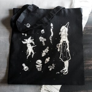 K&ouml;nnte beinhalten: Schwarze Tragetasche mit einer wei&szlig;en Illustration einer Hexe, die eine Blume h&auml;lt, umgeben von verschiedenen Pflanzen, Pilzen und einer Schlange. Der Text "STAG" befindet sich in der rechten unteren Ecke.