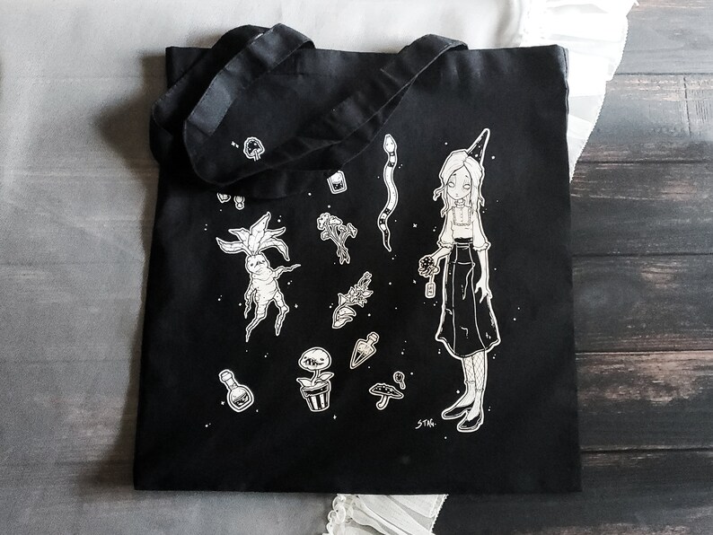 K&ouml;nnte beinhalten: Schwarze Tragetasche mit einer wei&szlig;en Illustration einer Hexe, die eine Blume h&auml;lt, umgeben von verschiedenen Pflanzen, Pilzen und einer Schlange. Der Text "STAG" befindet sich in der rechten unteren Ecke.