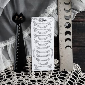 May include: A black cat-shaped bookmark and a ruler with inch measurements are next to a sticker sheet with bone and butterfly illustrations. The sticker sheet has the text "Tiny Bones" and "FB-1307". The ruler is silver and has a moon phase design.