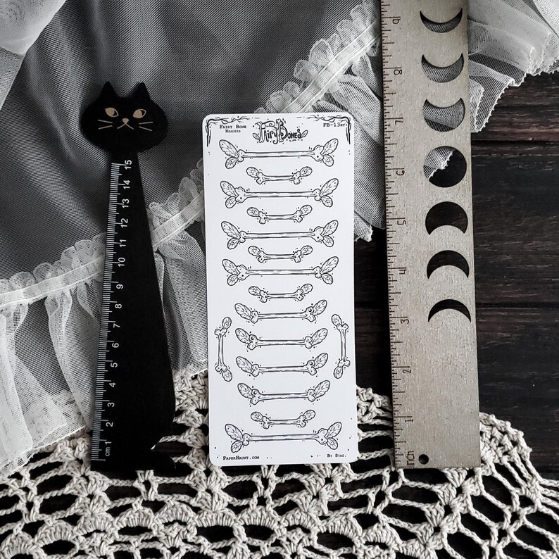 May include: A black cat-shaped bookmark and a ruler with inch measurements are next to a sticker sheet with bone and butterfly illustrations. The sticker sheet has the text "Tiny Bones" and "FB-1307". The ruler is silver and has a moon phase design.