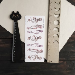 May include: A sheet of stickers featuring five illustrations of skeleton mermaids with pink and purple hues. The text "Fishbones: Mermaids" and "Art by Sta" are printed on the sheet.