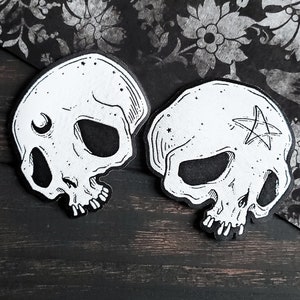 May include: Two black and white skull illustrations with details, one with a crescent moon and the other with a pentagram.