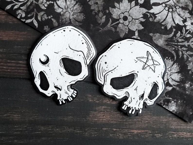 Op de afbeelding: Twee zwart-witte schedel-illustraties met details, een met een halve maan en de andere met een pentagram.