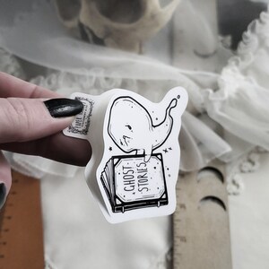 May include: A white sticker with a black and white illustration of a ghost whale sitting on a book that says "Ghost Stories".