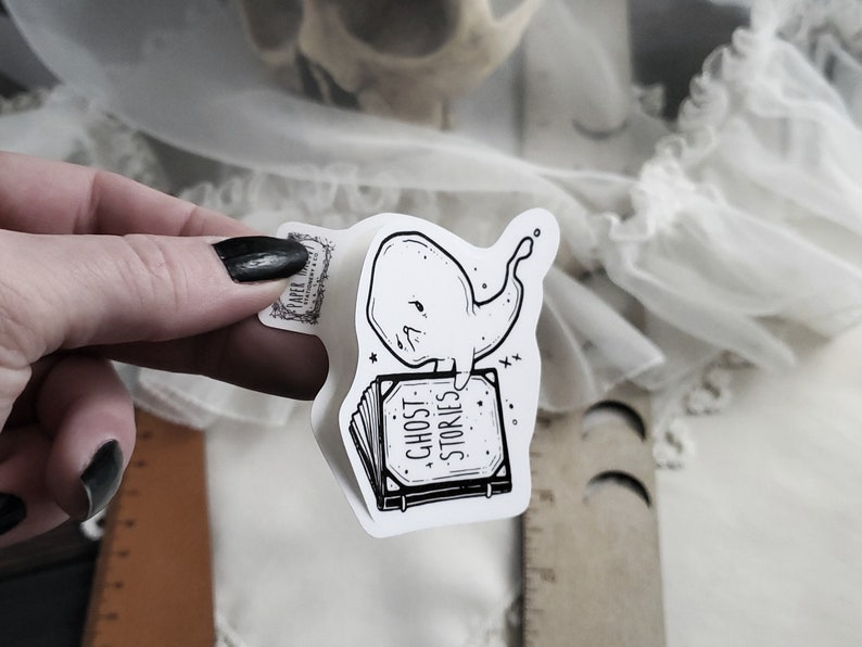 May include: A white sticker with a black and white illustration of a ghost whale sitting on a book that says "Ghost Stories".