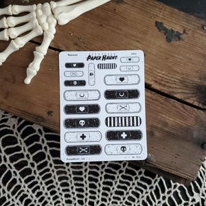 May include: A sheet of black and white illustrated band-aid stickers with various designs, including skulls, crosses, hearts, and crescent moons. The sheet is titled "Paper Haunt" and has the text "Band-Aids" at the top.